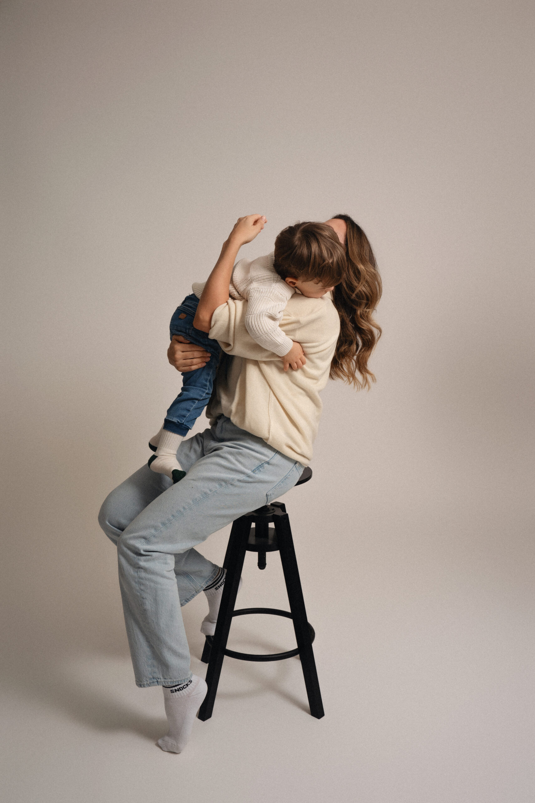 Mutter sitzt auf einem Hocker und hält ihr Kind liebevoll im Arm bei einem natürlichen Studio Fotoshooting