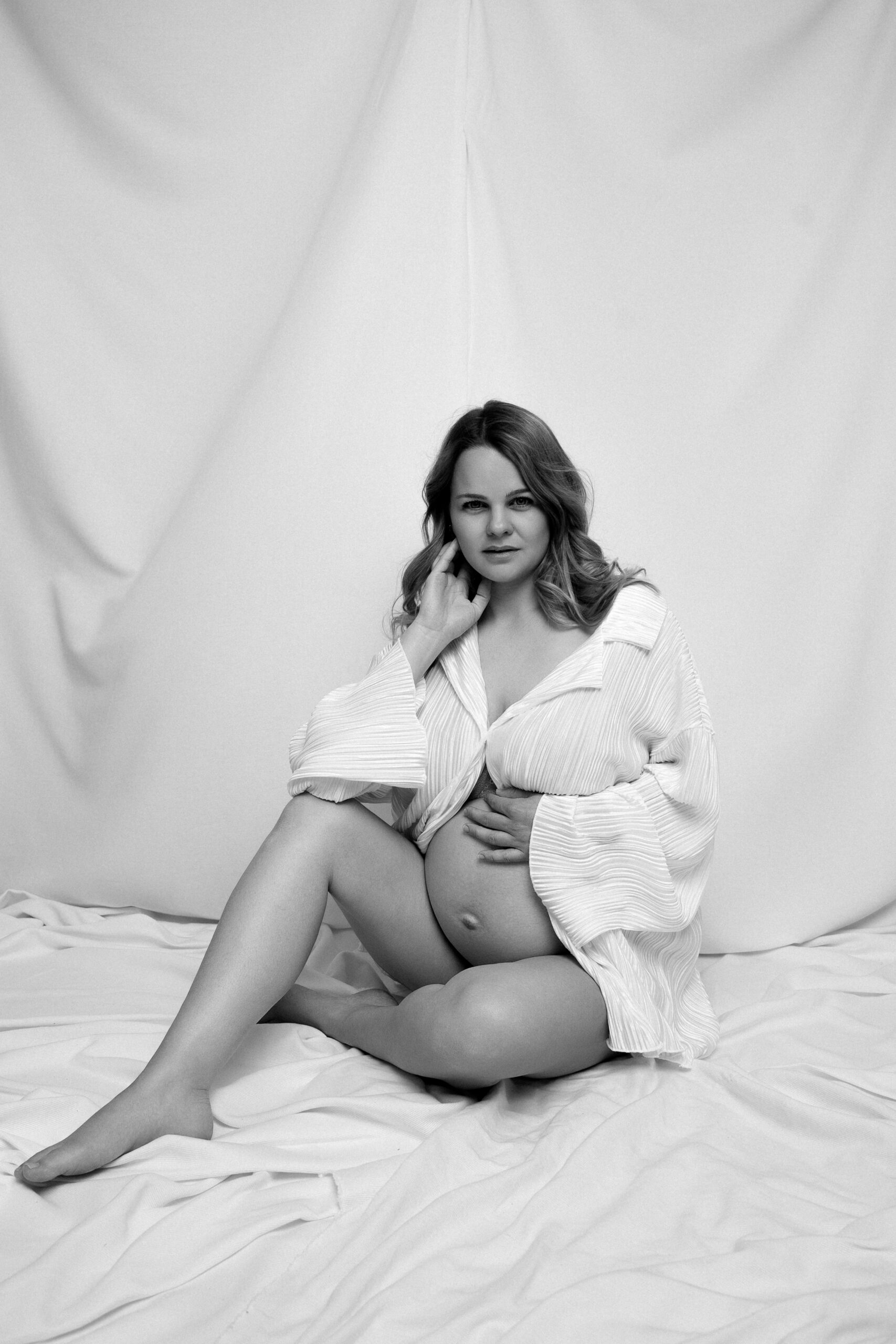 Schwangere Frau sitzt im Studio und hält ihren Babybauch bei einem natürlichen Schwarz-Weiß Fotoshooting