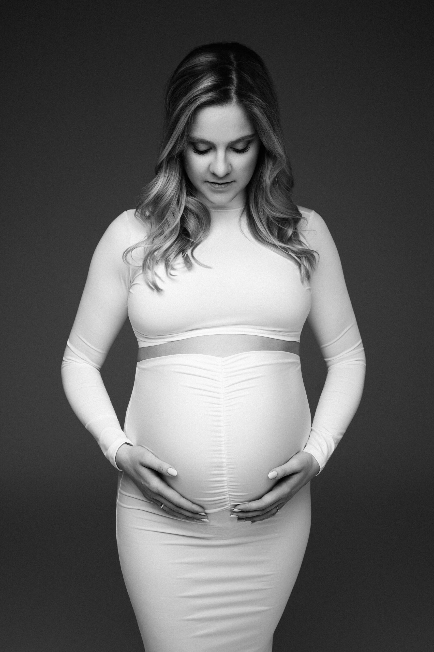 Werdende Mutter blickt auf ihren Babybauch in einem weißen Kleid bei einem Schwangerschaftsshooting in Ulm.