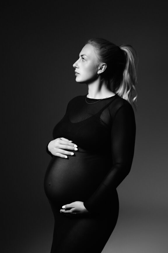 Schwangere Frau bei einem eleganten Schwarzweiß-Babybauchshooting in Ulm im Fotostudio mit schwarzem Kleid und Seitenprofil.